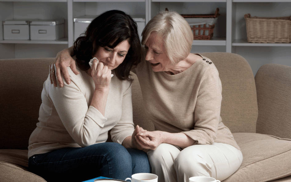 Two women sitting in a living room wearing beige coloured clothing. The woman one the left looks upset and is clutching a tissue while the woman on the right comforts her.
