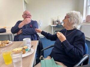 Two of our service users, an elderly man and woman chatting and laughing with snacks in front of them.