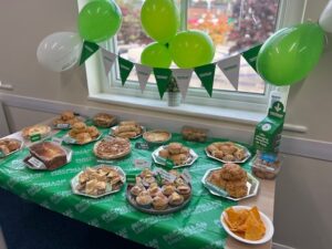 Spread of sweet treats on a green tablecloth and green decorations celebrating Macmillan Coffee Morning and fundraise.